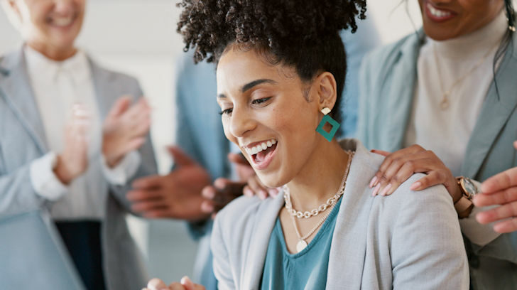 A woman smiling being congratulated by her coworkers around her.